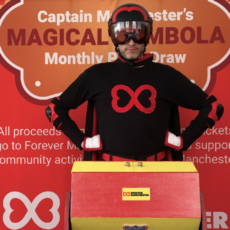 Captain Manchester stands in front of the tombola drum and Forever Manchester backdrop to announce the winners