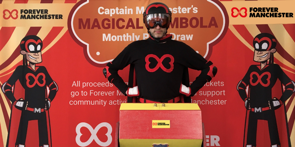 Captain Manchester stands in front of the tombola drum and Forever Manchester backdrop to announce the winners